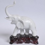 Elefante em porcelana branca esmaltada, base vazada em material sintético, medindo 9X11,5 cm. Uma das presas faltando pedaço.