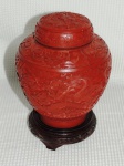 Potiche bojudo confeccionado em laca chinesa na cor vermelha ricamente entalhado com flores e acanto e tapa decorada com dragão em relevo. acompanha base em madeira. Med.: 22 cm.